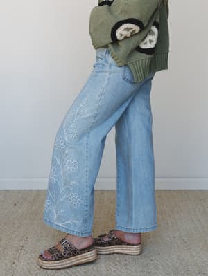 A side-profile shot showing wide-leg light wash jeans with delicate white floral embroidery climbing the leg, paired with an olive green peace sign sweater and leopard print platform sandals — the kind of outfit that looks effortless but definitely w