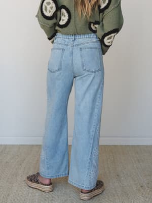 A back-view shot of wide-leg light wash jeans with subtle floral embroidery along the hem, paired with an olive crochet peace-sign sweater and leopard print platform espadrilles — the kind of outfit that feels effortless but clearly thought out.