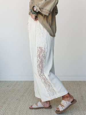A side-profile shot highlighting wide-leg ivory pants with a sheer lace inset panel running the length of the leg, styled with an olive embroidered jacket, stacked beaded bracelets, and double-buckle sandals — the kind of outfit that feels intentiona