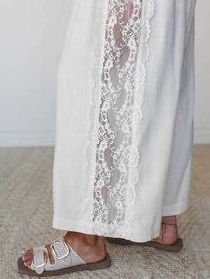 A close-up detail shot of wide-leg ivory pants with a sweeping vertical lace inset panel, styled with neutral double-buckle sandals on a woven rug — soft, romantic, and very much the kind of thing you'd want to wear to an outdoor wedding.