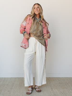 A layered boho look built around wide-leg ivory linen-look pants with lace side panels, styled with a graphic tee, pink floral-trim denim jacket, and stacked turquoise jewelry — the kind of outfit that feels totally pulled together without trying.