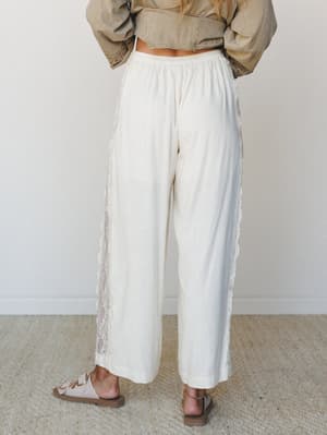 A back-facing shot of wide-leg ivory linen pants with lace side panel detailing, styled with a cropped olive jacket and neutral sandals — easy, romantic, and effortlessly put-together.