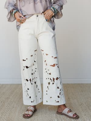 A cropped mid-shot showing wide-leg cream jeans with dramatic all-over eyelet cutwork, styled with a flowy lavender paisley blouse, stacked beaded bracelets, and neutral buckle sandals — the kind of outfit that looks effortless but absolutely has a p