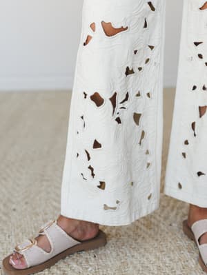 A close-up detail shot of wide-leg cream eyelet pants with intricate floral cutwork, paired with blush buckle sandals on a woven jute rug — the kind of outfit that makes a Tuesday feel intentional.