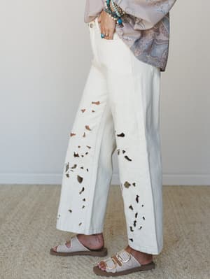 A side-angle shot of wide-leg cream jeans with scattered eyelet cutouts and subtle floral embroidery, styled with a paisley blouse, stacked turquoise bracelets, and neutral buckle sandals — effortlessly boho with a handcrafted feel.