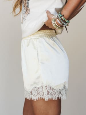 A close side-body shot of soft lemon satin shorts with a wide smocked waistband and pretty lace trim hem, styled with a white graphic tee and a stack of turquoise and pearl bracelets — the kind of outfit that feels equally at home on a lazy morning o