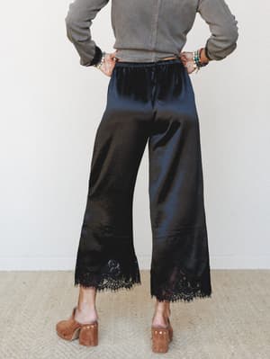 Shot from behind, a model wears wide-leg black satin trousers with a delicate lace hem, paired with a washed olive cropped top, stacked boho bracelets, and tan block-heel clogs — the kind of outfit that works harder than it looks.