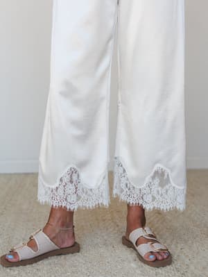 A close-up detail shot of wide-leg satin pants in champagne white, finished with a scalloped floral lace hem — styled casually with double-buckle sandals and a delicate ankle bracelet.