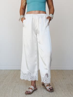 Wide-leg pants in a soft champagne white with floral lace trim at the hem, styled with a teal crop top, beaded bracelets, and buckle sandals — easy and expressive, like a beach vacation that somehow also works for brunch.