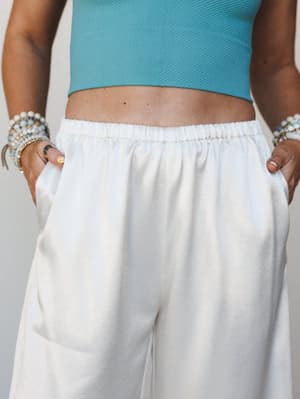 A close-up torso shot of wide-leg white pants with an elastic waistband, styled with a cropped teal textured top and layered beaded bracelets — relaxed and easy with just enough personality.