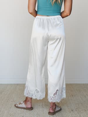 Shot from behind, a model in wide-leg satin pants with a sweeping lace hem trim pairs them with a teal crop top and neutral sandals — soft, romantic, and the kind of pants that make a Saturday feel intentional.