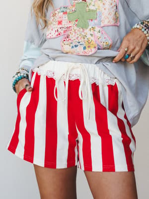 Bold red and cream wide-stripe shorts with a lace-trim waistband and drawstring tie, styled with a patchwork appliqué sweatshirt and stacked boho jewelry for a playful, personality-forward look.