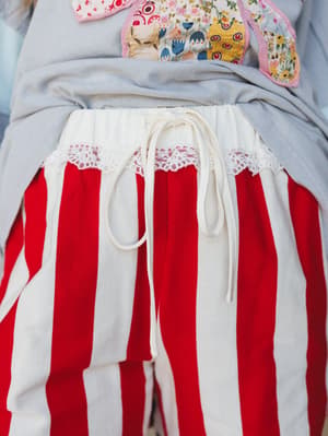 A close-up detail shot of bold red-and-cream striped shorts with a drawstring waist and delicate lace trim at the hip, layered over a soft grey top with playful patchwork appliqué peeking in at the top.