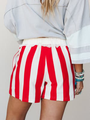 A back-view shot showing bold red and white striped shorts with a lace-trimmed waistband, layered under a pale blue oversized top and styled with stacked turquoise and beaded bracelets — casual and a little nostalgic, like a summer Saturday with nowh