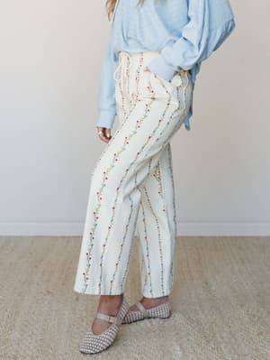 A cropped mid-shot of wide-leg cream pants covered in delicate vertical floral vine embroidery, styled casually with a powder blue sweatshirt and woven mary jane flats — the kind of outfit that feels effortless but clearly thought about.