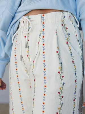 A close-up of cream wide-leg pants covered in vertical rows of colorful floral vine embroidery — sunny yellows, reds, blues, and greens — paired with a soft blue cropped top, giving the whole look a garden-party-meets-everyday-magic feel.