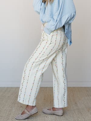 Wide-leg cream pants covered in delicate vertical floral vine embroidery are styled with a soft powder blue oversized top and woven ballet flats — the kind of outfit that feels like a slow Sunday morning with somewhere worth going.