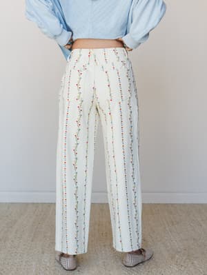A back-view shot of wide-leg cream pants covered in delicate vertical floral vine embroidery in red, blue, and yellow — paired with a light blue top and woven flats for an effortlessly pretty, garden-ready look.