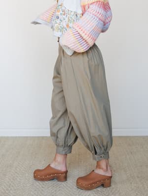 A side-angle shot of voluminous olive barrel pants with elasticated cuffs, paired with a colorful striped knit sweater, a floral underlayer peeking through, and warm brown studded clogs — the kind of outfit that looks effortless but definitely took t