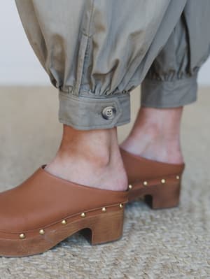 A close-up detail shot of gathered, button-cuffed olive barrel pants paired with cognac leather clogs on a wood sole — earthy, intentional, and the kind of combo that makes you want to rearrange your whole closet.