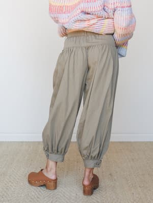 Shot from behind, a model pairs relaxed olive barrel pants — gathered at the waist and elasticated at the ankle — with a chunky multicolor sweater and warm brown clogs, giving the whole thing an effortless, intentional-weekend kind of energy.