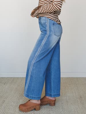 A side-profile shot of wide-leg cropped denim jeans with contrast seam detailing, styled with a brown striped long-sleeve top and cognac wood clogs — a lived-in, 70s-leaning combo that feels effortless without trying.