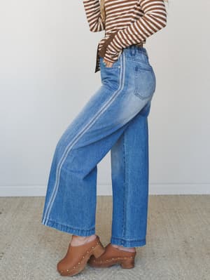 A side-profile shot of wide-leg denim jeans in medium wash, styled with a brown striped long-sleeve top and tan clogs — very much a '70s-leaning, lived-in look that feels effortless without trying.