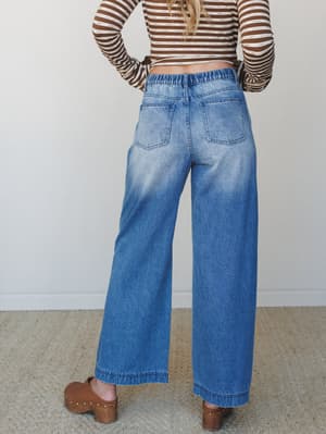 Shot from behind, this look pairs wide-leg medium-wash jeans — with a relaxed elastic waistband and patch pockets — with a brown striped crop top and wooden clogs, giving off that effortless 70s-leaning everyday energy.