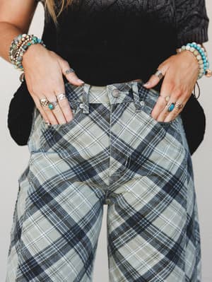 A close-up detail shot of plaid wide-leg pants in faded sage and navy, styled with a dark cable-knit crop and layered with turquoise beaded bracelets and stacked silver rings — the kind of outfit that looks effortless but definitely wasn't accidental