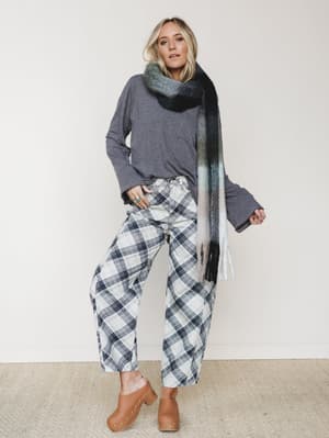 A relaxed full-body studio shot with a cozy, collected feel — plaid wide-leg pants paired with an oversized gray long-sleeve and a gradient fringe scarf, finished with tan clogs for that effortless fall-into-winter vibe.