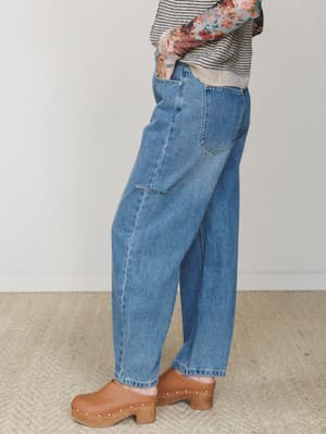 A side-profile shot of relaxed barrel-leg jeans in medium wash denim, styled with a striped sweater, floral top layer, and tan studded clogs — the kind of outfit that looks effortless but was definitely thought about.