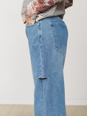 A close-up side shot of wide-leg distressed denim jeans in a classic medium wash, styled with a striped top and layered floral scarf — the kind of lived-in pair that goes with everything and gets better every wash.