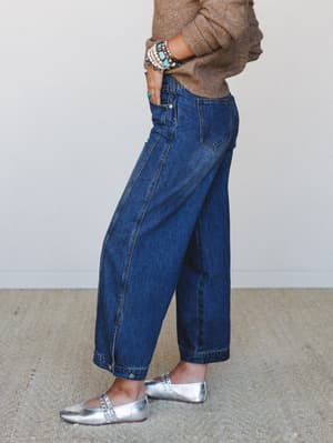 A side-profile shot of wide-leg cropped denim jeans styled with a camel knit sweater, silver Mary Jane flats, and layered turquoise and pearl bracelets — the kind of effortless, put-together look that feels like it happened naturally.