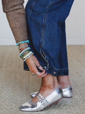 A close-up of a model adjusting her wide-leg denim jeans near the hem, styled with silver metallic mary jane flats and a stacked wrist full of turquoise, stone, and silver beaded bracelets — the kind of detail shot that makes you want to copy the who