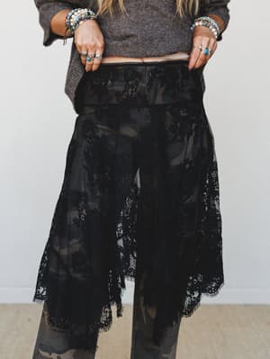 A close-cropped studio shot of a sheer black lace overlay skirt styled over camo pants with a cropped grey sweater, the model's stacked beaded bracelets and silver rings adding a layered boho finish.