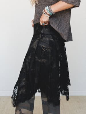 A black floral lace midi overlay skirt with an asymmetric handkerchief hem is styled over camo leggings and paired with a charcoal brushed sweater — the mix of tough, soft, and romantic all at once, accessorized with a stack of boho bracelets and a t