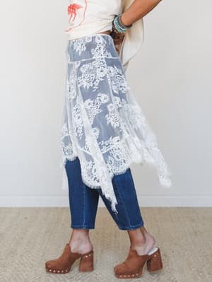 A sheer floral lace overlay skirt worn over dark denim jeans, styled with a graphic tee and brown studded clogs — that effortless mix of soft and lived-in that feels completely intentional.