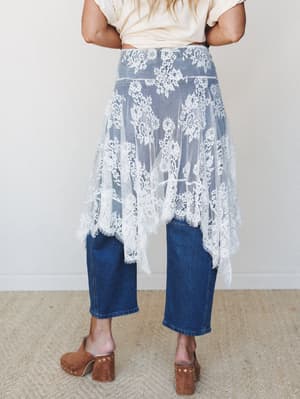 A sheer floral lace overlay skirt worn over dark wash cropped wide-leg jeans, styled with a cream tee and brown suede clogs — the kind of unexpected layering that makes a basic outfit feel like a whole personality.
