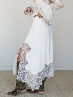 A side-profile shot of a cream midi skirt with dramatic lace hem and side slit, paired with a sheer lace bell-sleeve top and leopard print cowboy boots — romantic boho energy with just enough edge.