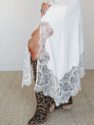 A cream midi skirt with sweeping lace trim and a thigh-high slit is styled with leopard print cowboy boots and a turquoise ring — the kind of outfit that feels equally at home at a backyard wedding or a country music festival.