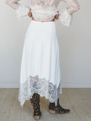 A cream midi skirt with a wide lace hem and handkerchief points is styled from the back with a sheer lace bell-sleeve crop top and leopard print cowboy boots — romantic and a little wild in the best way.