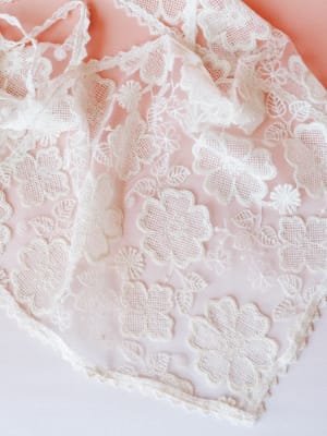A close-up flat lay of a cream lace layering skirt with delicate floral appliqué and scalloped edges, laid against a soft blush-to-white gradient background — the kind of piece that makes you want to plan a whole outfit around it.