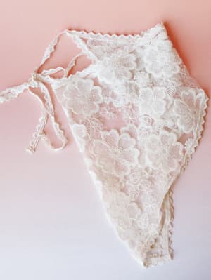 A delicate cream lace triangle skirt laid flat against a soft blush-to-white gradient background — all floral appliqué and tie closure, the kind of thing you'd layer over a swimsuit or slip dress and feel completely yourself.