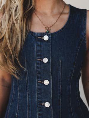 A close-up torso shot of a fitted dark denim vest top with silver snap buttons and seamed paneling, styled with a delicate gold charm necklace for an effortless, vintage-leaning feel.