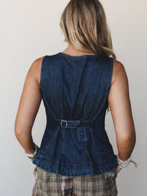 A back-view shot of a dark denim belted vest top with a fitted peplum silhouette, styled with plaid shorts and layered bracelets against a clean studio backdrop — vintage-meets-everyday in the best way.