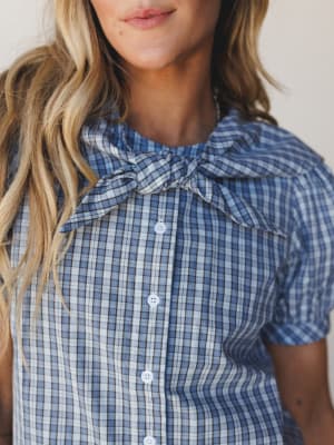 A close-up of a plaid button-front blouse in dusty blue with a self-tie bow collar and short sleeves — the kind of top that feels vintage-sweet without trying too hard.