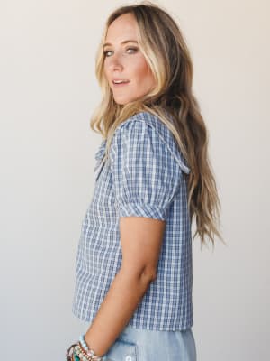 A side-profile shot of a model in a dusty blue plaid blouse with puff sleeves and a bow neckline, styled with light wash jeans and stacked boho bracelets — easy, warm, and a little nostalgic.
