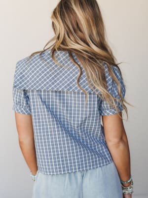 A back-view shot of a cropped plaid blouse in dusty blue with puff sleeves and a yoke detail, styled with light chambray pants and turquoise bead bracelets — casual and collected with a quiet vintage feel.