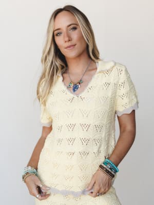 A soft, sun-washed crochet knit top in buttery yellow with a relaxed v-neck collar and layered boho jewelry — the kind of top that makes a Tuesday feel like a beach town.