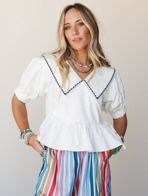 A breezy white peplum top with an oversized sailor collar and navy ric-rac trim, styled with a multicolor striped skirt and layered beaded necklaces — the kind of outfit that makes a Tuesday feel like a summer trip somewhere.